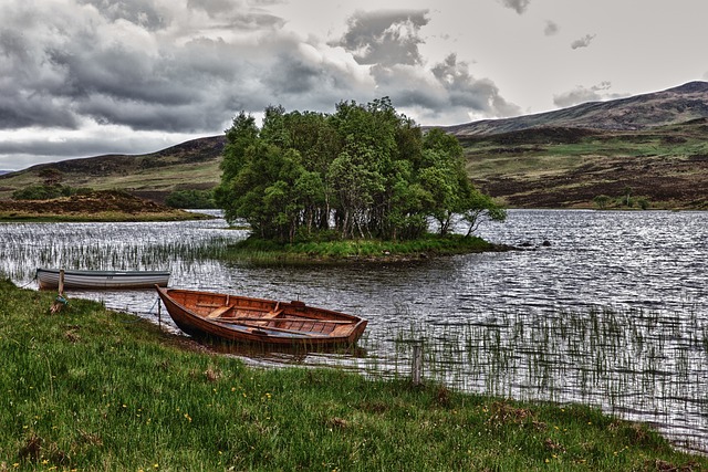 Two wooden rowboats rest on the grassy shore of a lake, with a small, tree-covered island in the center under a dramatic, cloudy Scottish sky.